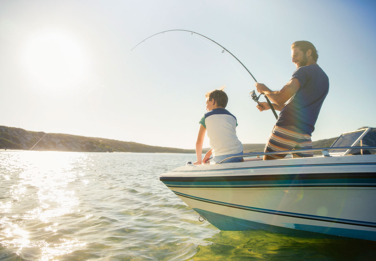 Older man reeling in fish with young boy watching from bow of boat.